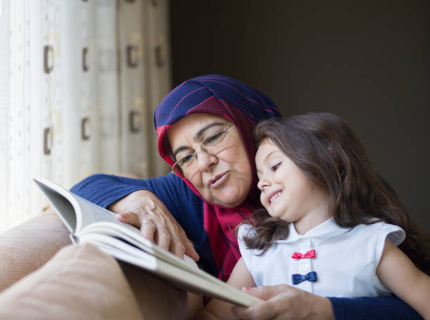 Grandmother wearing a head scarf reading book with granddaughter